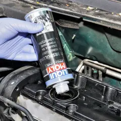 Ręka w niebieskiej rękawiczce wlewa Liqui Moly płukanie silnika do otworu w pokrywie zaworów.