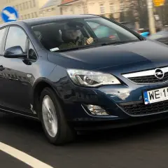 Ciemnoniebieski Opel Astra J 1.6 benzyna w ruchu miejskim. Kierowca w czapce prowadzi auto.