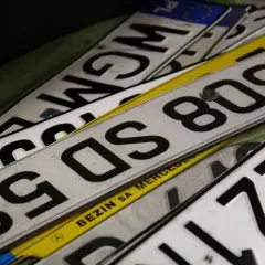 Stos **tablic rejestracyjnych państw** w różnych kolorach i z numerami. Widać fragmenty z PL, F, a także napisy "BEZIN SA MERCEDES".
