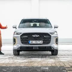 Mężczyzna w pomarańczowej kurtce pokazuje kciuki w górę obok nowego, szarego SUV-a. To jeden z chińskich aut, który zyskuje na popularności.