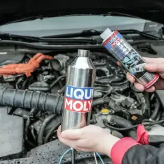 Dwie ręce trzymają produkty Liqui Moly: metalową butelkę i puszkę z napisem "Catalytic System Cleaner". W tle widać silnik samochodu.