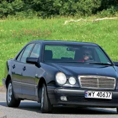 Czarny Mercedes W210 z silnikiem om606 jedzie po drodze, za nim zielone, kwieciste wzgórze.