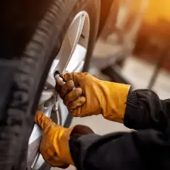 Ręce w żółtych rękawicach odkręcają śruby koła, przygotowując się do wymiany, jak wyjąć koło zapasowe Citroen Berlingo.