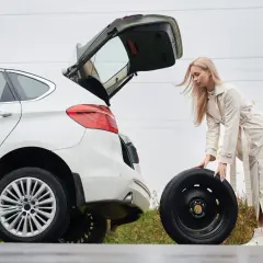 Kobieta wyjmuje koło zapasowe z bagażnika białego auta. W tle widać drogę i jesienne drzewa. To może być pomocne, jak zdjąć koło zapasowe Citroen Jumper.