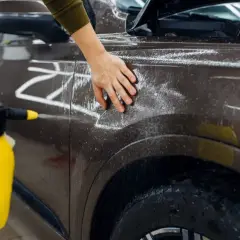 Ręka dotyka brązowej karoserii samochodu pokrytej pianą z żółtego opryskiwacza.