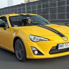 Żółte coupé Toyota GT86 z czarnymi pasami na masce jedzie po drodze.