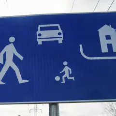 Niebieski znak drogowy ze schematycznymi symbolami: pieszy, samochód, dziecko grające w piłkę, dom.