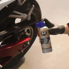 Ręka w czarnej rękawiczce trzyma puszkę smaru do łańcucha K2. Motocykl w tle.