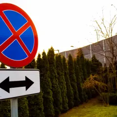 Znak zakaz zatrzymywania z tabliczką wskazującą kierunki. W tle budynek i żywopłot.