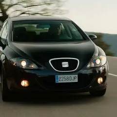 Czarny Seat Leon 2 jedzie po krętej drodze wśród drzew.