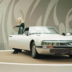 Elegancka blondynka otwiera drzwi białego Citroëna SM na wystawie. Obok stoją mężczyźni w garniturach.