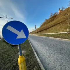 Niebieski znak drogowy ze strzałką w prawo na tle zielonego wzgórza i nieba.