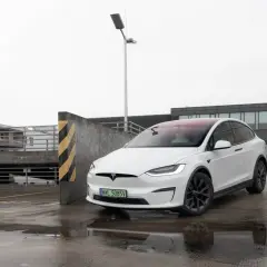 Biały Tesla Model X na mokrym parkingu, z widokiem na budynki i latarnię.