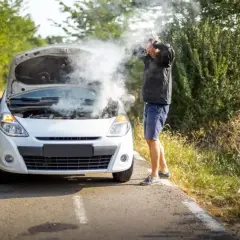 Mężczyzna z rozpaczą patrzy na biały samochód z otwartą maską, z której bucha dym. Rozbiegany diesel na poboczu drogi.