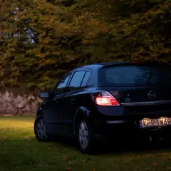 Czarny Opel Astra H 1.6 benzyna zaparkowany na trawie, w tle jesienne drzewa.