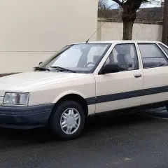 Jasny beżowy sedan Renault 21 z lat 80. stoi na mokrej drodze.