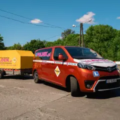 Pomarańczowy van z przyczepą szkoleniową "POLDEK.pl" na placu manewrowym.