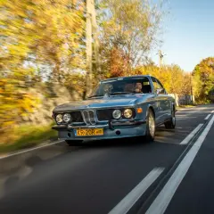 Błękitne BMW E9 z kierowcą jedzie jesienną drogą, otoczone kolorowymi drzewami.