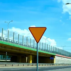 Pomarańczowy znak drogowy "ustąp pierwszeństwa" na tle wiaduktu i błękitnego nieba.