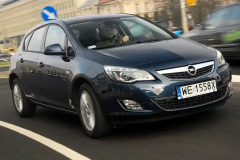 Ciemnoniebieski Opel Astra J 1.6 benzyna w ruchu miejskim. Kierowca w czapce prowadzi auto.
