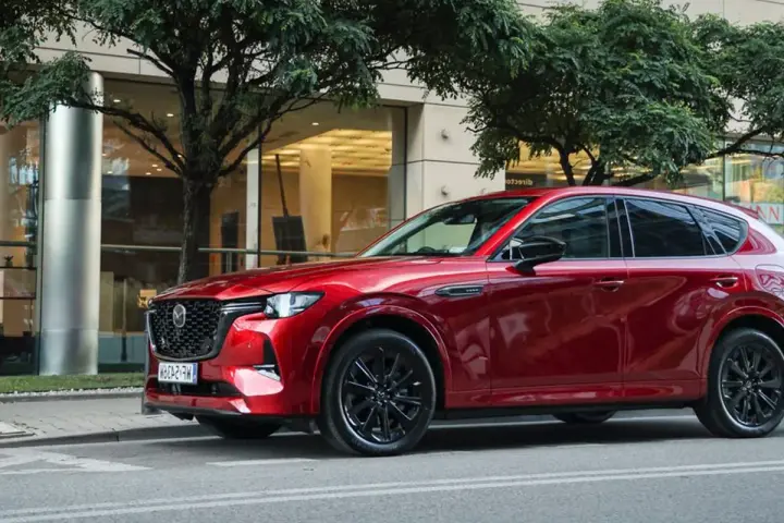 Lśniąca, czerwona Mazda CX-60 3.3 diesel na tle nowoczesnego budynku i drzew.