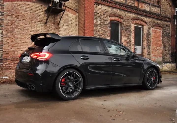 Czarny Mercedes-AMG A45s z dużym spojlerem i czerwonymi zaciskami hamulcowymi stoi przed starą ceglaną kamienicą.
