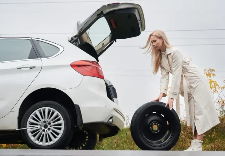 Kobieta wyjmuje koło zapasowe z bagażnika białego auta. W tle widać drogę i jesienne drzewa. To może być pomocne, jak zdjąć koło zapasowe Citroen Jumper.