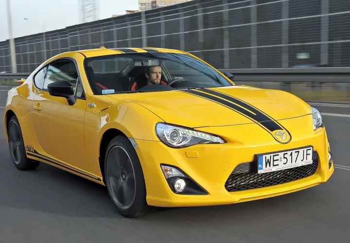 Żółte coupé Toyota GT86 z czarnymi pasami na masce jedzie po drodze.
