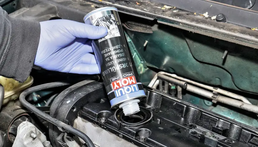 Ręka w niebieskiej rękawiczce wlewa Liqui Moly płukanie silnika do otworu w pokrywie zaworów.