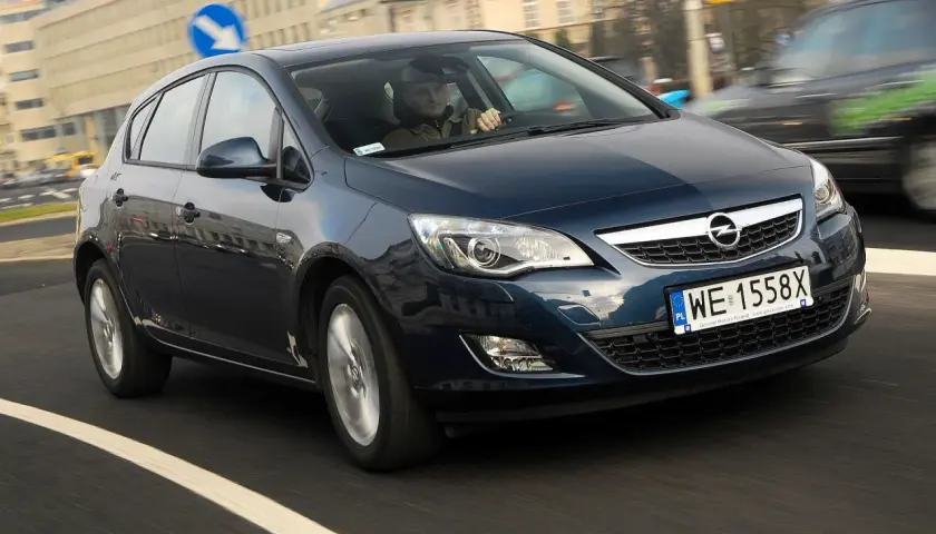 Ciemnoniebieski Opel Astra J 1.6 benzyna w ruchu miejskim. Kierowca w czapce prowadzi auto.