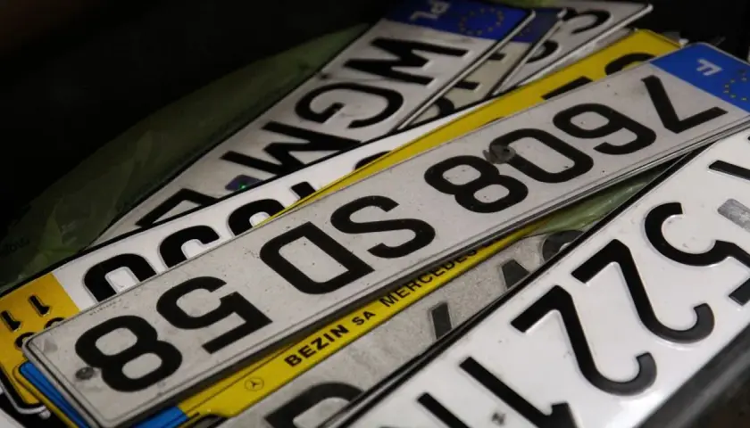 Stos **tablic rejestracyjnych państw** w różnych kolorach i z numerami. Widać fragmenty z PL, F, a także napisy "BEZIN SA MERCEDES".
