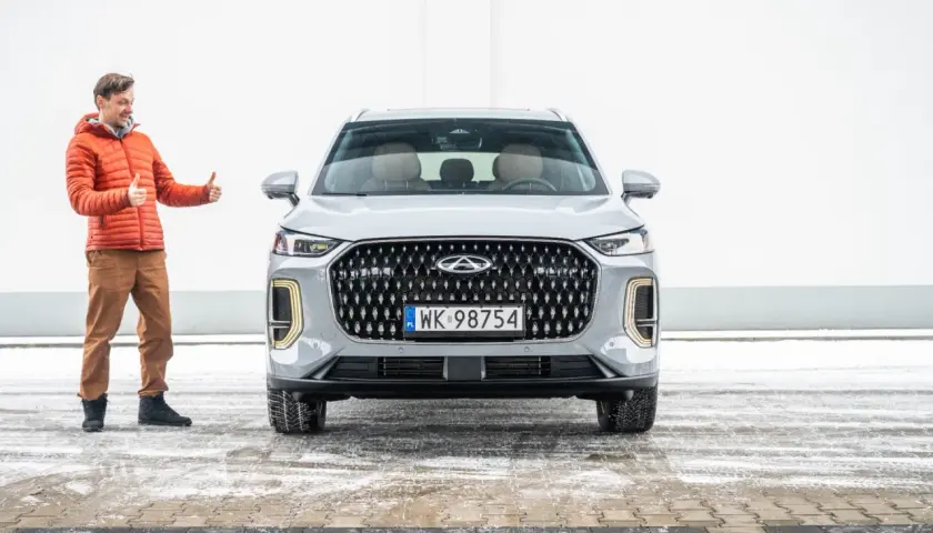 Mężczyzna w pomarańczowej kurtce pokazuje kciuki w górę obok nowego, szarego SUV-a. To jeden z chińskich aut, który zyskuje na popularności.