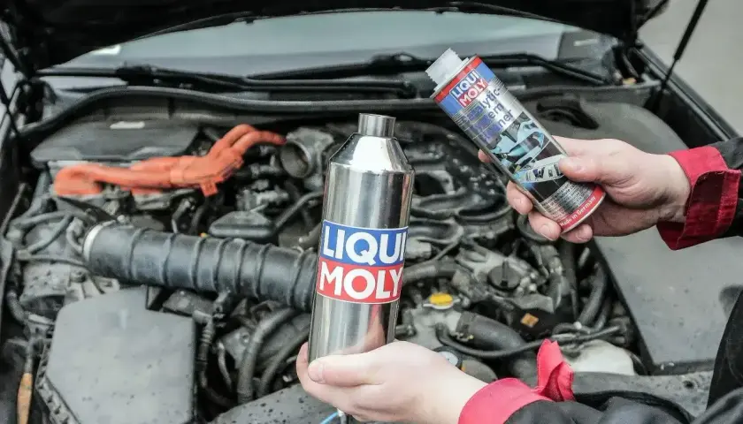 Dwie ręce trzymają produkty Liqui Moly: metalową butelkę i puszkę z napisem "Catalytic System Cleaner". W tle widać silnik samochodu.