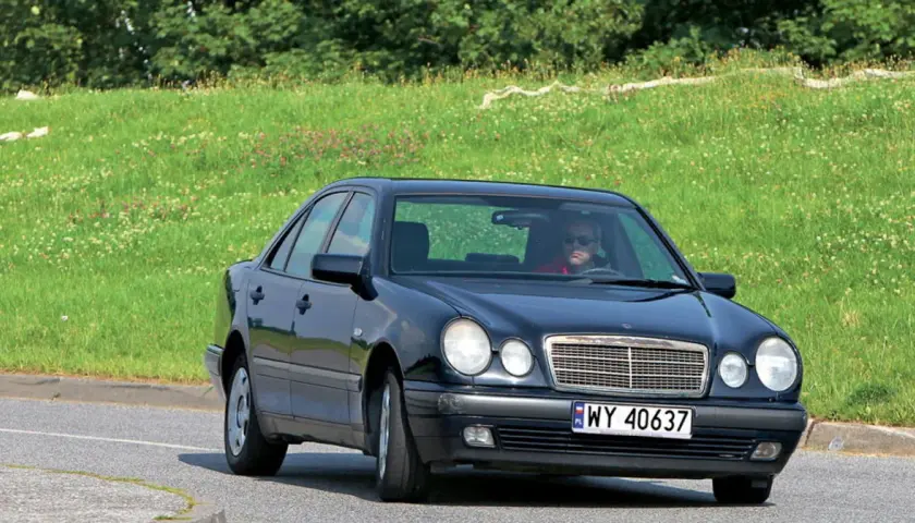 Czarny Mercedes W210 z silnikiem om606 jedzie po drodze, za nim zielone, kwieciste wzgórze.