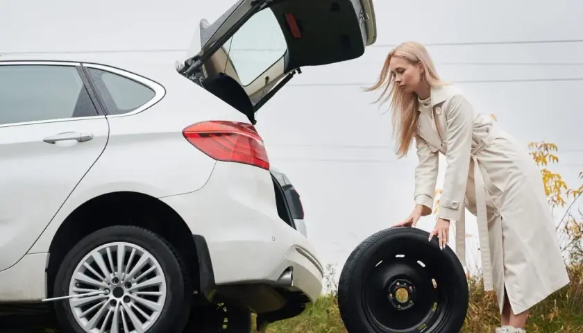 Kobieta wyjmuje koło zapasowe z bagażnika białego auta. W tle widać drogę i jesienne drzewa. To może być pomocne, jak zdjąć koło zapasowe Citroen Jumper.