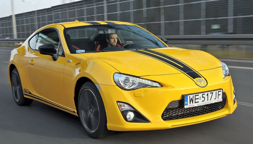 Żółte coupé Toyota GT86 z czarnymi pasami na masce jedzie po drodze.