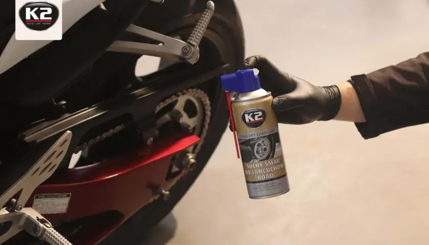Ręka w czarnej rękawiczce trzyma puszkę smaru do łańcucha K2. Motocykl w tle.