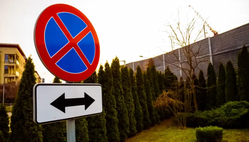 Znak zakaz zatrzymywania z tabliczką wskazującą kierunki. W tle budynek i żywopłot.