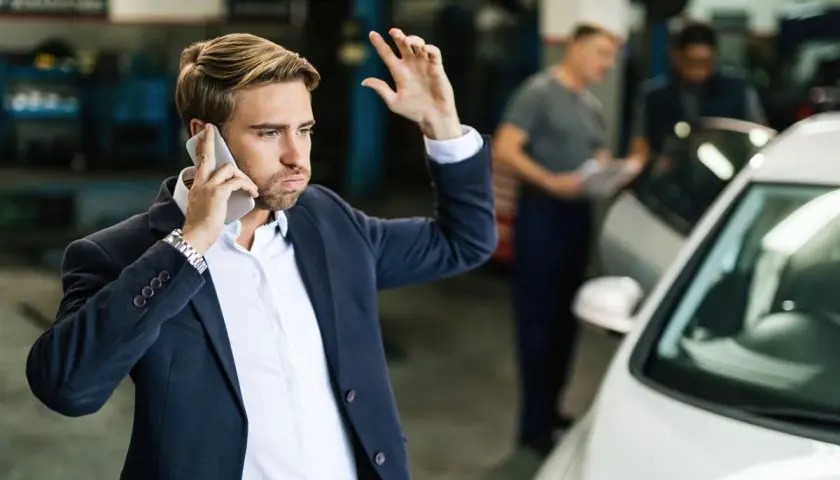 Mężczyzna w garniturze rozmawia przez telefon, gestykulując ręką, obok białego samochodu w warsztacie.