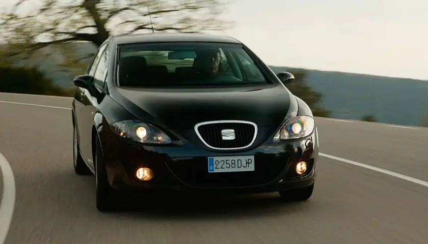 Czarny Seat Leon 2 jedzie po krętej drodze wśród drzew.