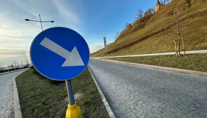 Niebieski znak drogowy ze strzałką w prawo na tle zielonego wzgórza i nieba.