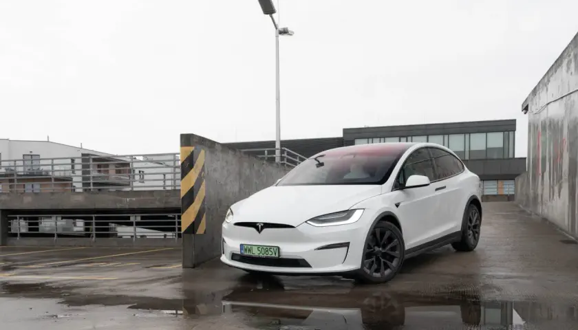 Biały Tesla Model X na mokrym parkingu, z widokiem na budynki i latarnię.