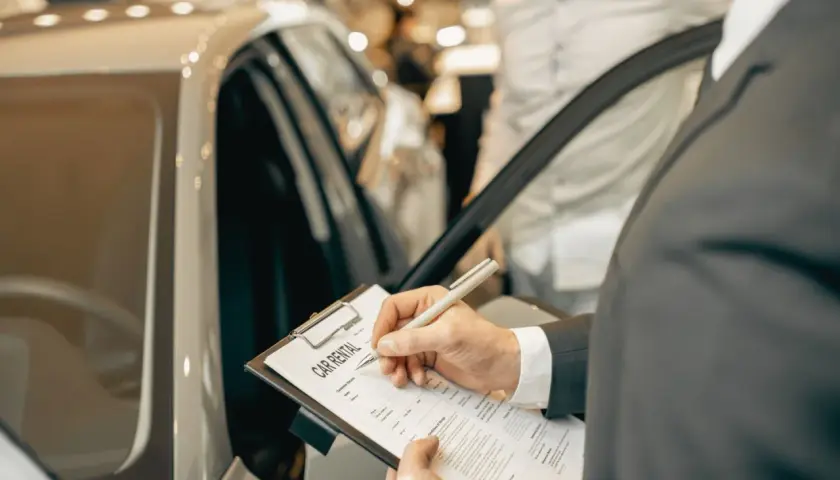 Mężczyzna w garniturze podpisuje dokument "CAR RENTAL" przy luksusowym samochodzie.