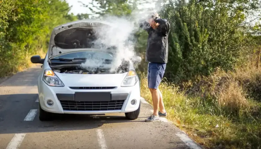 Mężczyzna z rozpaczą patrzy na biały samochód z otwartą maską, z której bucha dym. Rozbiegany diesel na poboczu drogi.