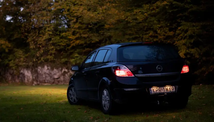 Czarny Opel Astra H 1.6 benzyna zaparkowany na trawie, w tle jesienne drzewa.