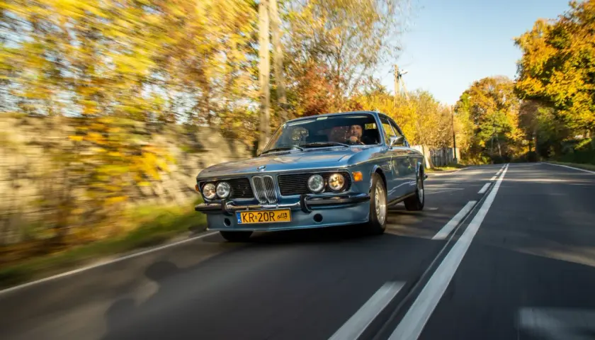 Błękitne BMW E9 z kierowcą jedzie jesienną drogą, otoczone kolorowymi drzewami.