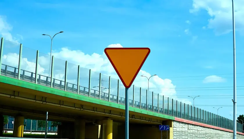 Pomarańczowy znak drogowy "ustąp pierwszeństwa" na tle wiaduktu i błękitnego nieba.