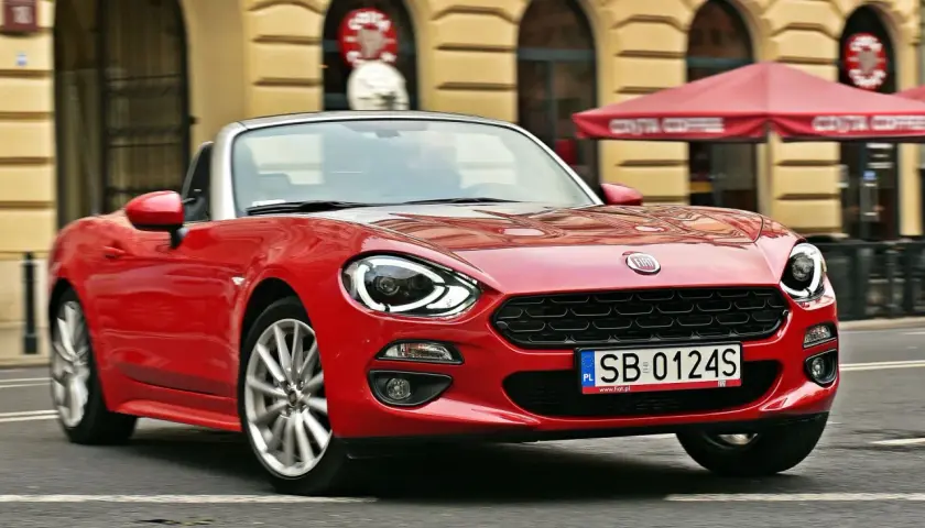 Czerwony Fiat 124 Spider z otwartym dachem sunie ulicą przed kawiarnią Costa Coffee.
