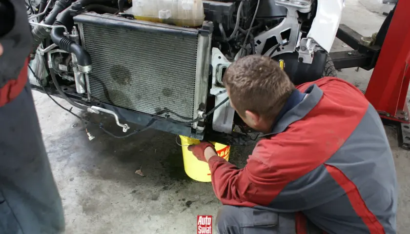 Mechanik w rękawiczkach nalewa środek do uszczelniania chłodnic do żółtego wiadra pod samochodem.