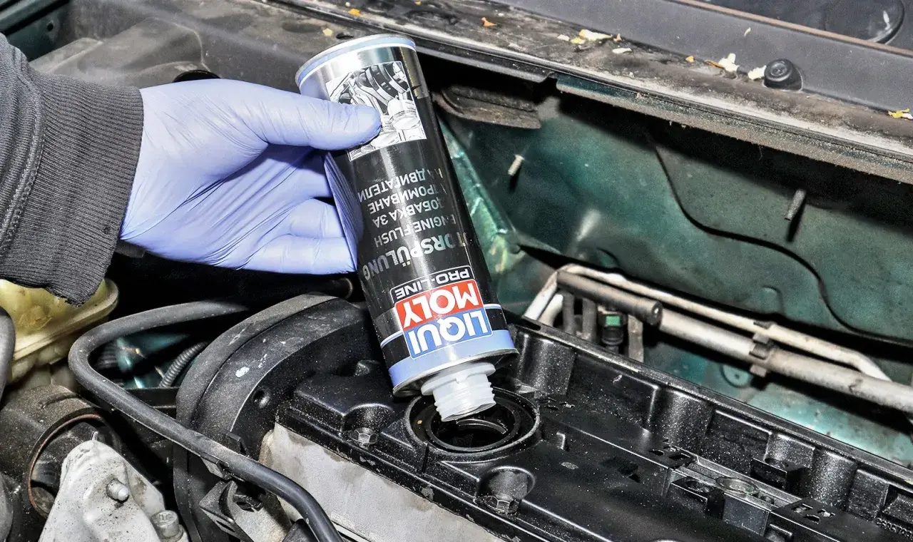 Ręka w niebieskiej rękawiczce wlewa Liqui Moly płukanie silnika do otworu w pokrywie zaworów.